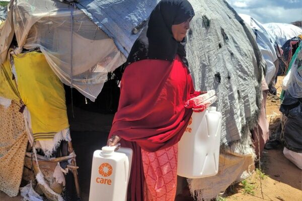 Dadaab Refugee Camp - outskirts - this is where the new arrivals, that have not been registered yet, settle. Fatuma, 60, is a new arrival at Dadaab. She arrived two months ago from Mogadishu in Somalia together with 12 members of her family. She is carrying two CARE jerricans and a package of soap bars, that have been distributed by CARE.

“There are no usable latrines for us, so we have to just go where we can. And we drink from the same water human and animal feces have been washed into. Some of my neighbors have gotten sick and are in the hospital,” describes Fatuma. In April this year, there have already been more than 1,000 confirmed cases of Cholera. Fatuma is not registered yet and lives in the outskirts with about 133.000 other new arrivals. “We came from Mogadishu, where my husband died in an explosion. There we had no security and no education. It was very hard. It took us eight days to come here. We saved the money for a truck to the border, then we walked the rest.” 
Eight family members fit in the small about 4m² makeshift hut made from sticks and cloth from old clothes that she and her children built themselves. “We have no supplies, everything here we either found or neighbors gave to us. It is not big enough for my whole family. Five of the boys sleep outside,” says Fatuma. The cloth and tarps that make up her home are not waterproof. The heavy rains completely flood her hut. Every night she sits inside with her three grandchildren under her clothes, and they pray, that the roof does not collapse on top of their heads. “It was painful to leave home, even talking about it now, my emotions come up. But where else should we go? At home in Somalia is nothing left, only fear and trauma.”
Living in the outskirts of the official camp is a challenge for Fatuma. Access to water, food, and basic items such as soap is limited. There are not enough resources for the many people in need. The distribution of soap and jerricans has been cancelled for three months in a row, because the resources are missing. CARE has stepped in and started distributing to new arrivals. “Without CARE we would have nothing. Now we at least have access to some drinking water, soap, and jerricans”, says Fatuma. However, the drinking water in the tanks built by CARE is not enough for everyone. Water deliveries are also delayed due to the current floods in Dadaab. “When there is no water in the tanks, we sometimes walk for 1.5 hours to go and find water. Then we roll the water back to our hut,” describes Fatuma. Rolling the water means that the round jerricans are put on the floor and rolled either by hand or by foot through mud, dirt, and feces. The clean drinking water easily contaminates this way and can cause serious illnesses. That is why CARE distributes rectangular jerricans, with an easy handle to carry, so the water stays clean and safe.

CARE supports the new arrivals with the supply and installation of micro water storage facilities which can hold to up to 5,000 liters of drinking water and distributes plastic jerricans, constructs communal latrines and supplies soap bars to all new arrival households residing in the outskirts. Additionally, women and girls are provided with menstrual hygiene dignity kits.